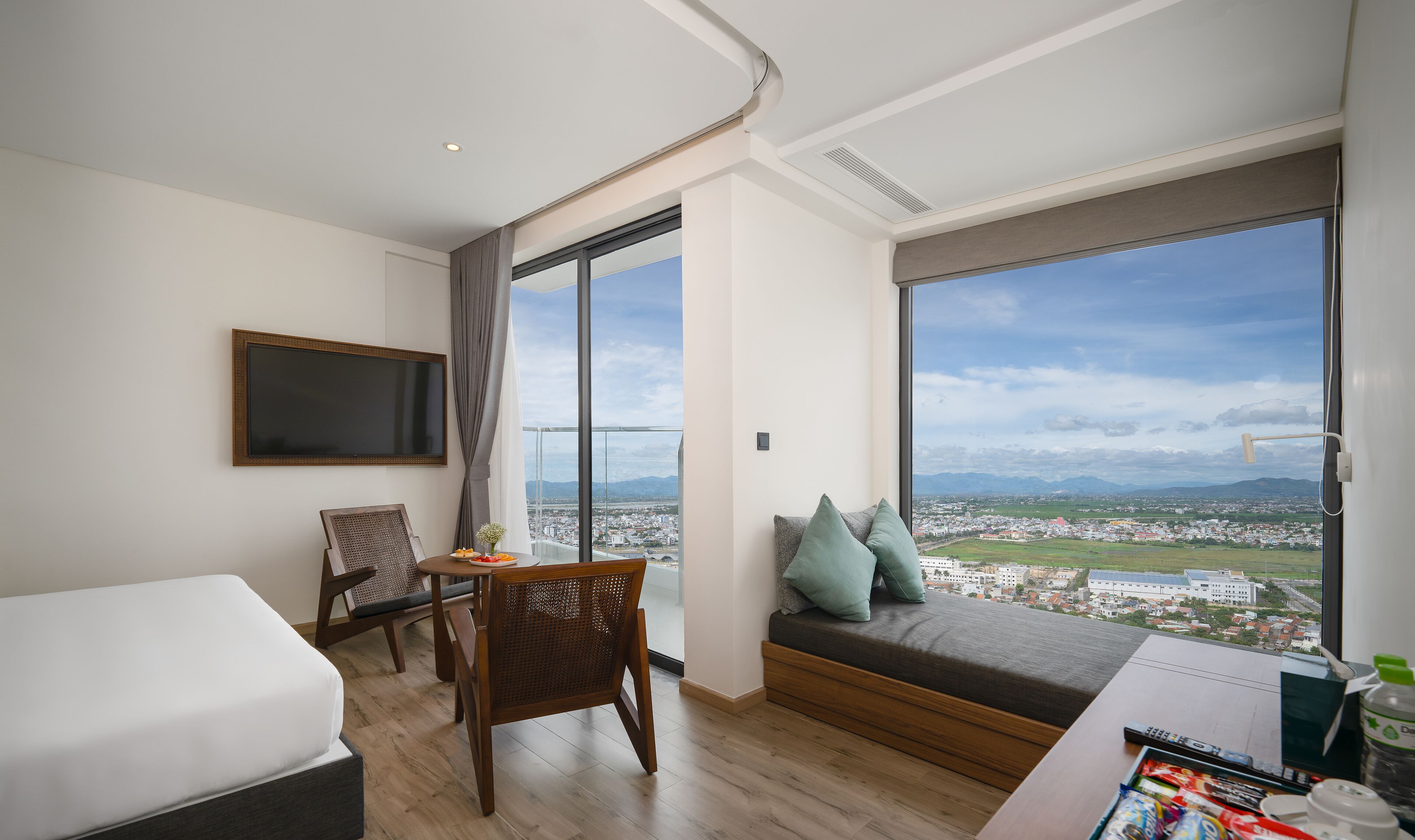 A hotel room with a bed, a desk, a chair, a large window with a city view, and a mounted TV.