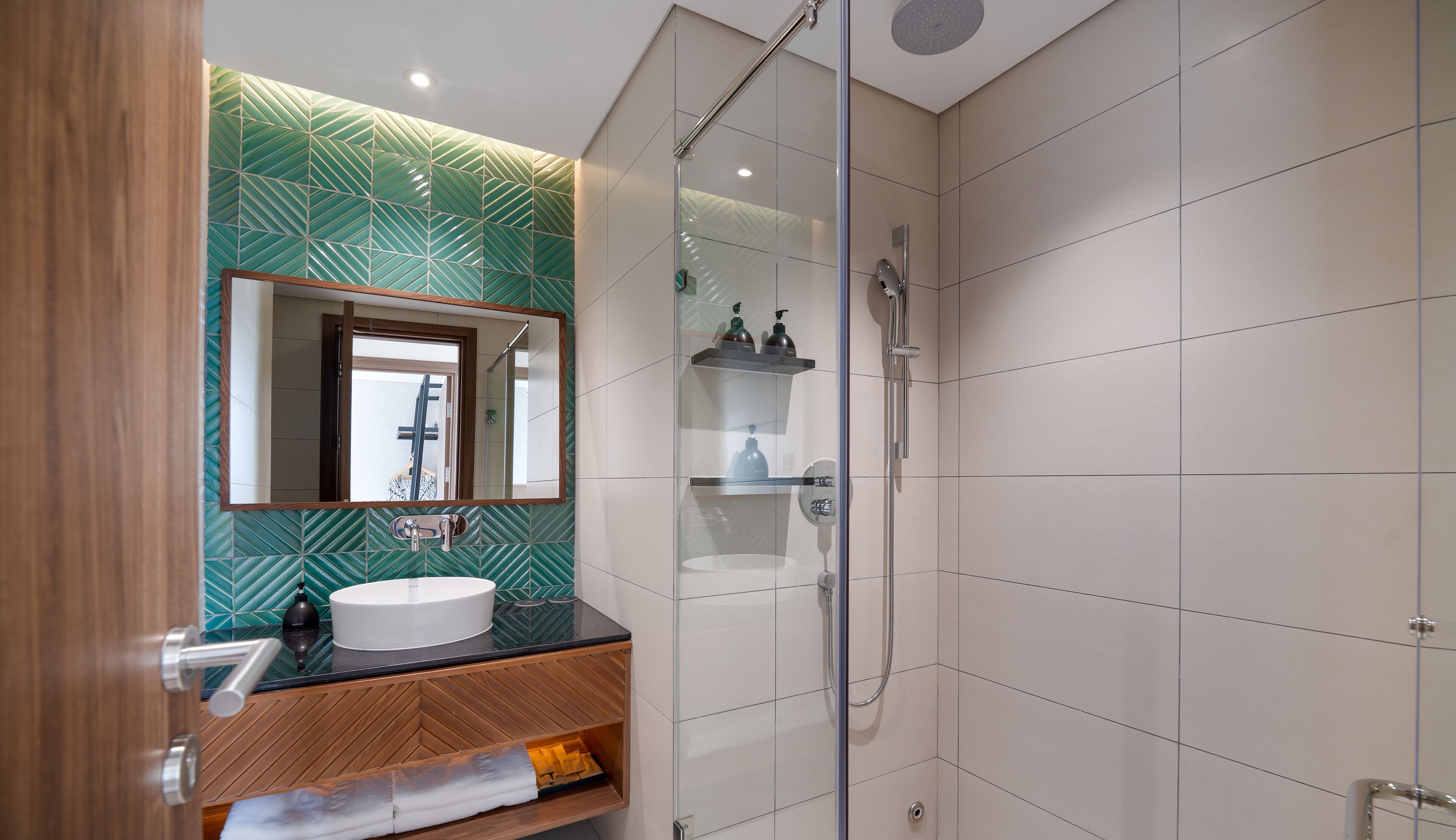 A modern bathroom with a glass shower enclosure, a wooden vanity, and a green patterned wall.