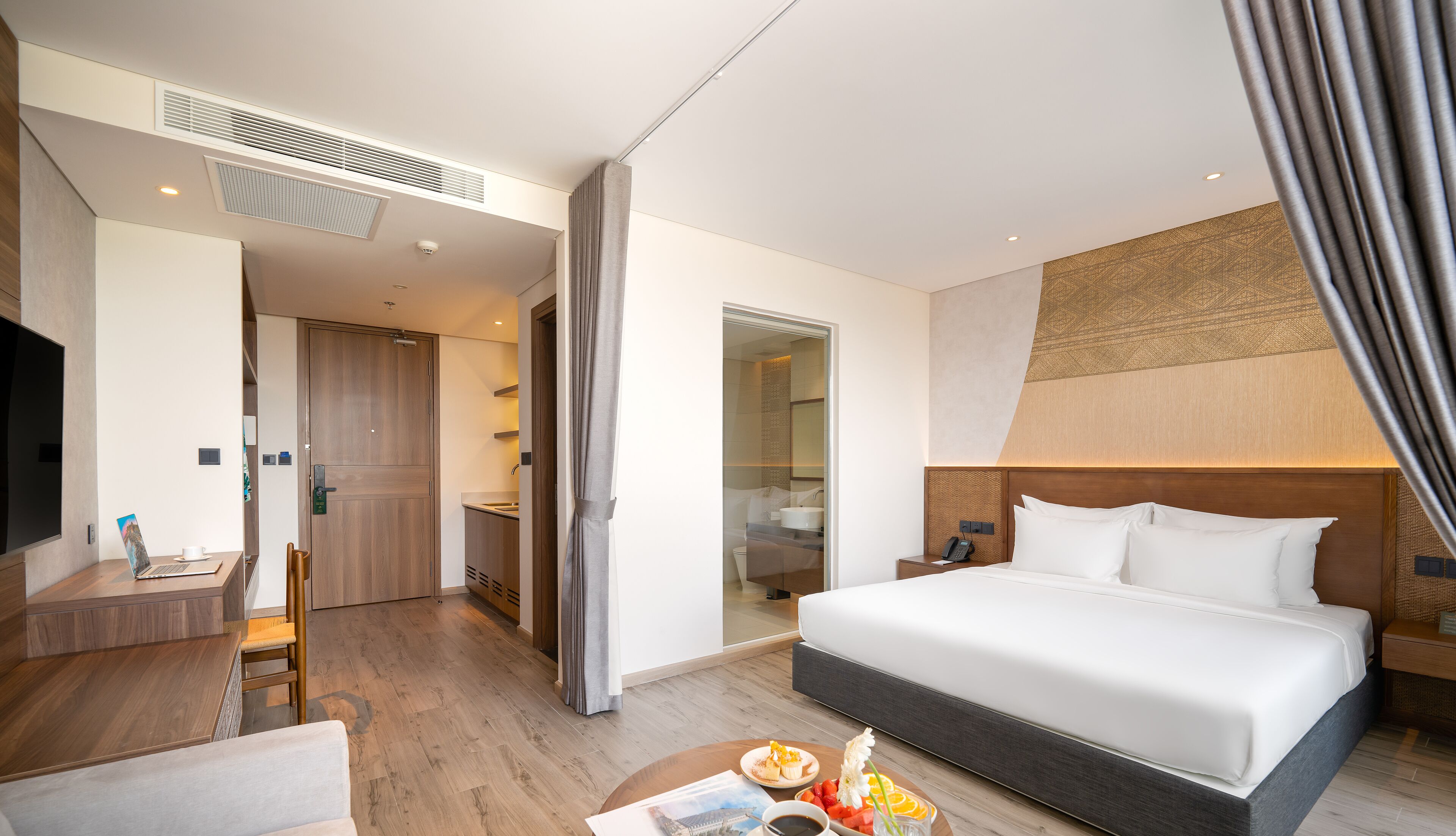 A modern hotel room with a large bed, a desk with a laptop, a bathroom visible through a glass door, and a dining area with a table set for two.