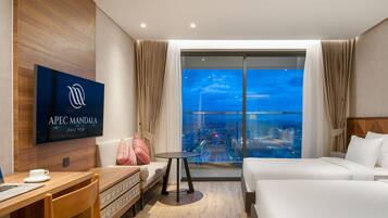 A modern hotel room with a large bed, a sofa, a small table, and a TV displaying the hotel name.