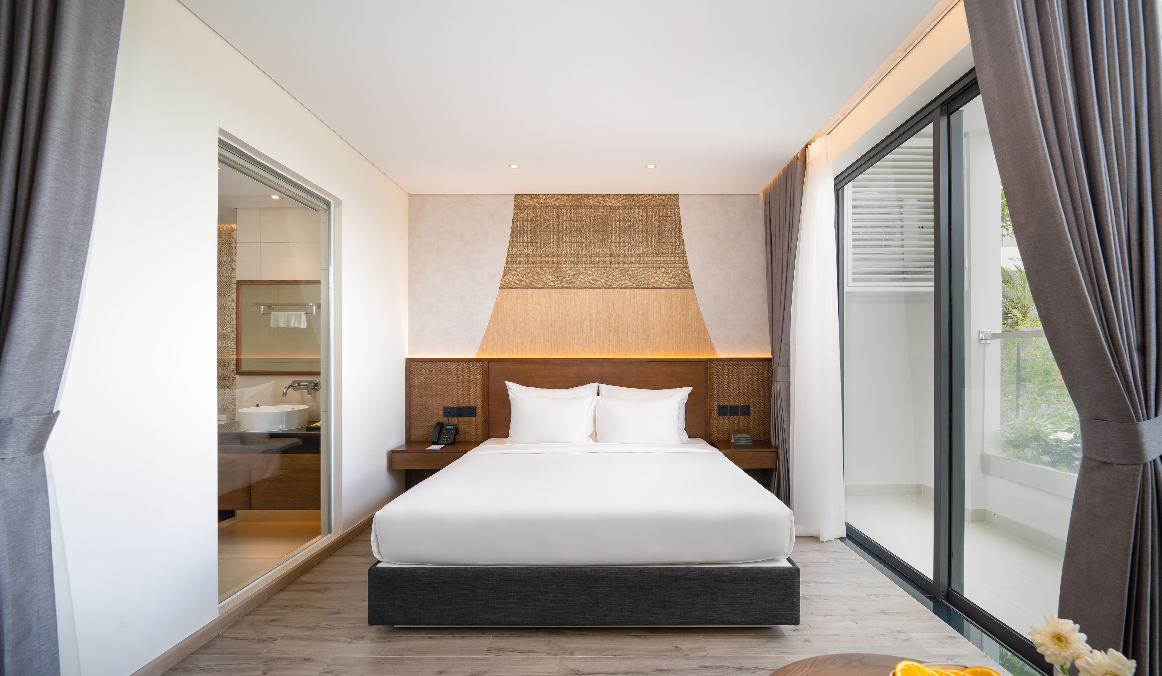 Una habitación de hotel moderna con una cama grande, un baño con lavabo y espejo, y vistas a través de un ventanal amplio.