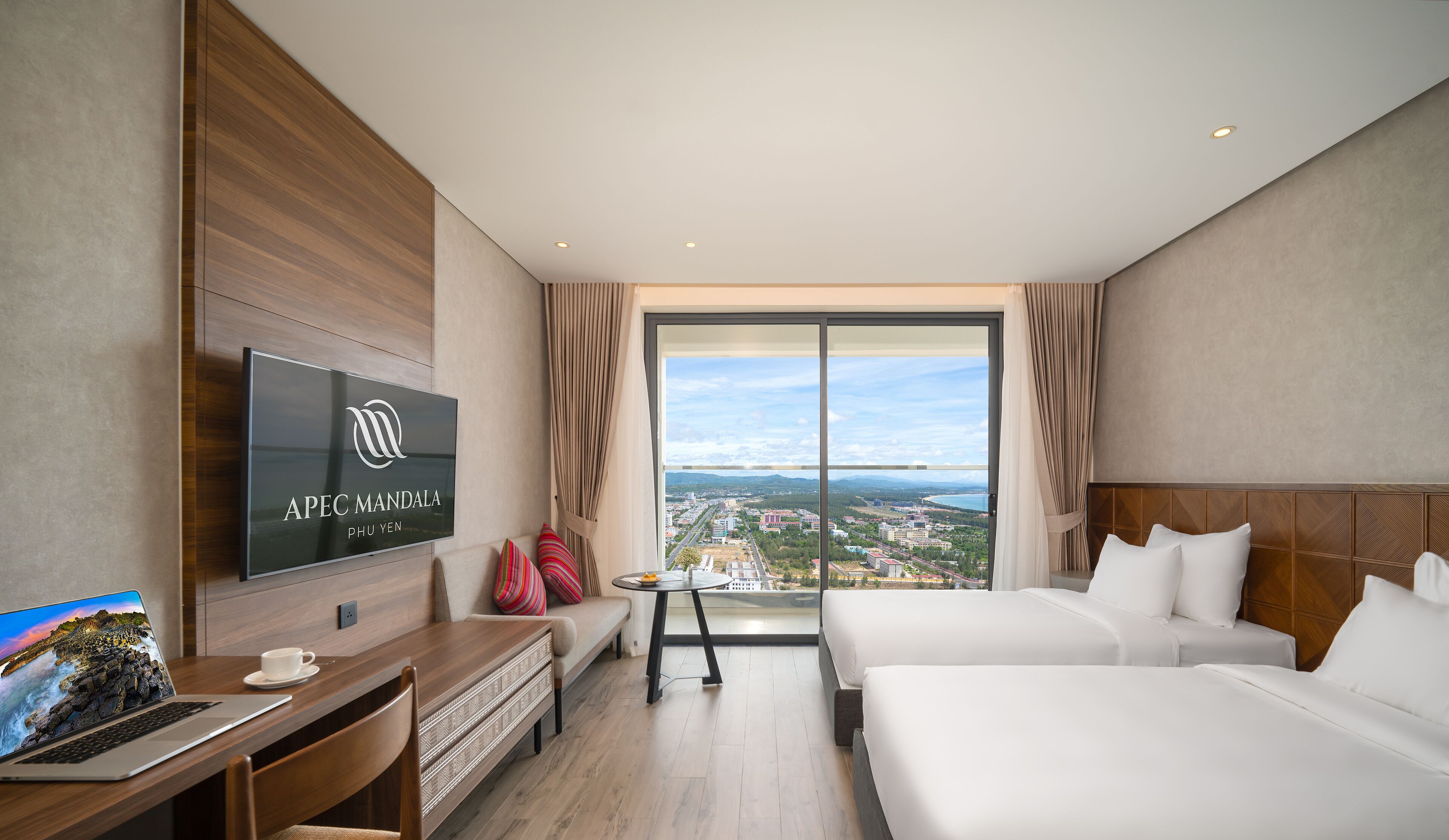 Habitación de hotel con televisor grande, escritorio con computadora portátil, cama y vista a la ciudad a través de un ventanal.