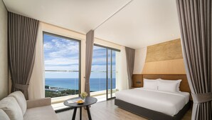 A modern hotel room with a large window offering a sea view, a bed, a sofa, and a small table with a vase of flowers. - Mandala Hotel & Spa Phu Yen - 24-hour stay (Tuy Hoa)