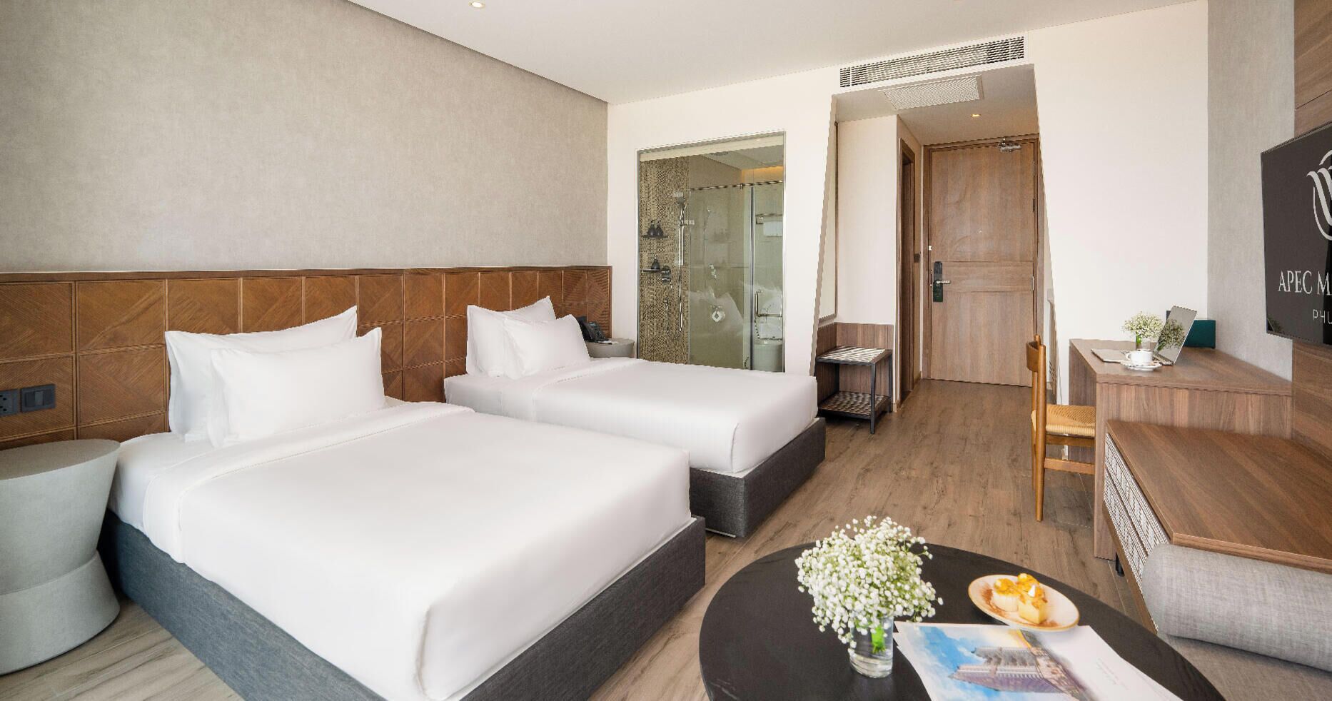 Une chambre d’hôtel avec deux lits, un bureau, une chaise, une petite table avec des fleurs et un espace douche.
