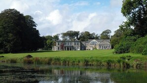 Un grand immeuble à étages, doté d’une façade symétrique, entouré d’une végétation luxuriante et bordé d’une étendue d’eau calme dans laquelle se reflète le bâtiment.