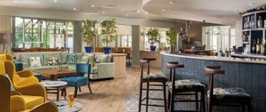 A modern bar area with a counter and stools, a seating area with sofas and chairs, and large windows allowing natural light.
