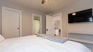 Ein Schlafzimmer mit einem Bett, einem Deckenventilator, einem an der Wand montierten Fernseher und einem durch eine offene Tür einsehbaren Badezimmer.