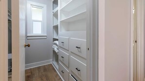 Ein schmaler begehbarer Kleiderschrank mit weißen Regalen und Schubladen, einem Fenster und einer Tür.