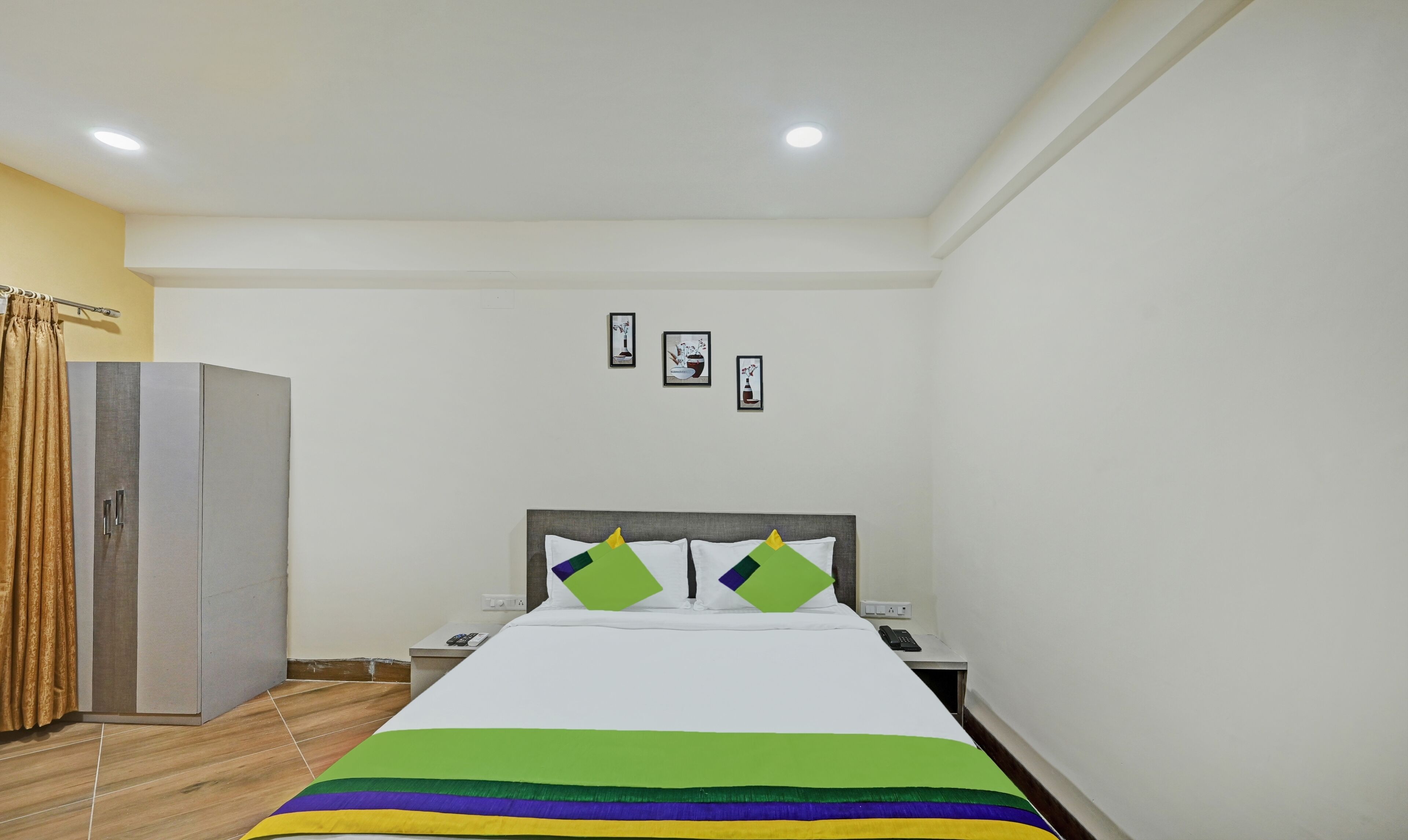 A hotel room with a bed, bedside tables, a wardrobe, and a wall with framed pictures.