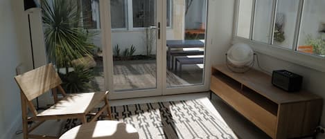 Una habitación moderna con una puerta corredera de vidrio que da a un patio, una silla de madera, una pequeña mesa redonda, un tapete estampado y un mueble consola de madera con un altavoz.
