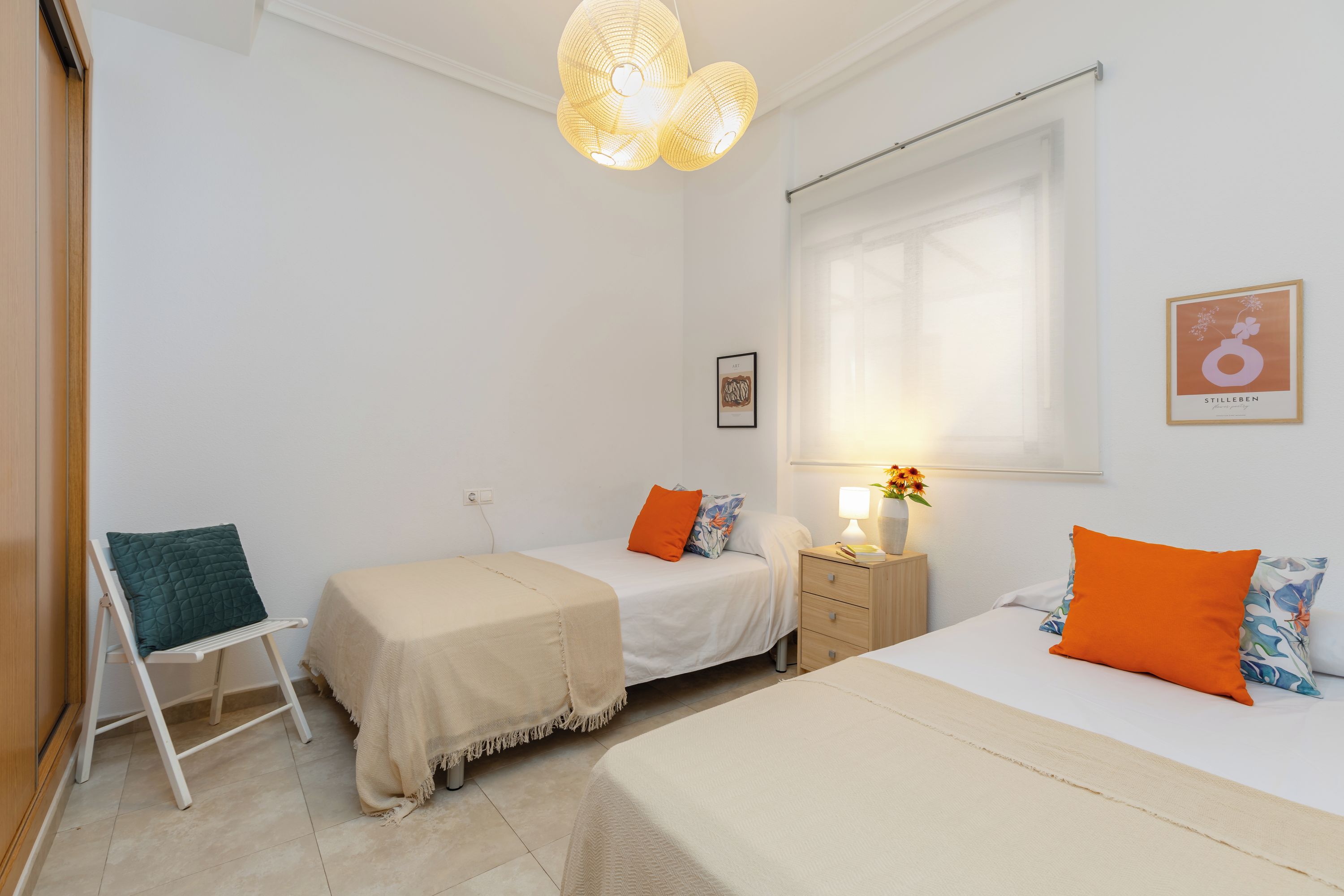 A bedroom with two beds, a nightstand, a chair, and a window with blinds.
