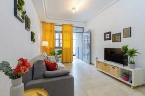 Una sala moderna con un sofá gris, un televisor sobre un mueble blanco y una planta en el mueble.