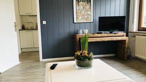 A modern living room with a white coffee table, a TV on a stand, a framed picture on the wall, and a kitchen in the background.