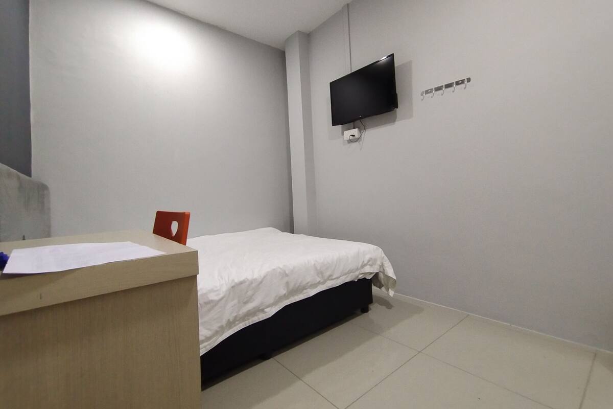 A bedroom with a bed, a desk with a chair, a flat-screen TV mounted on the wall, and a coat rack.