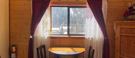 Interior de una cabaña de madera con una mesa redonda y dos sillas, una ventana con cortinas y un armario de madera.