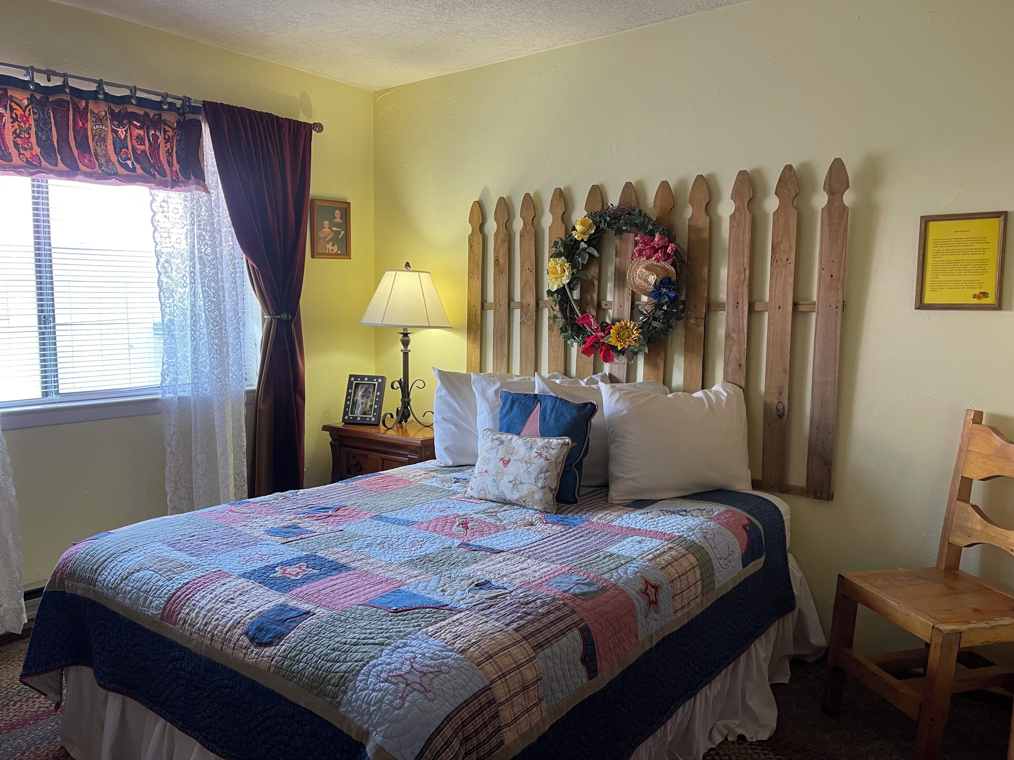 A bedroom with a bed, a wooden headboard, a nightstand with a lamp, a chair, a window with curtains, and a wall with a wreath and a framed picture.