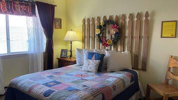 A bedroom with a bed, a wooden headboard, a nightstand with a lamp, a chair, a window with curtains, and a wall with a wreath and a framed picture.