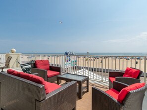A balcony with wicker furniture and red cushions, overlooking a beach with people and a clear sky.
