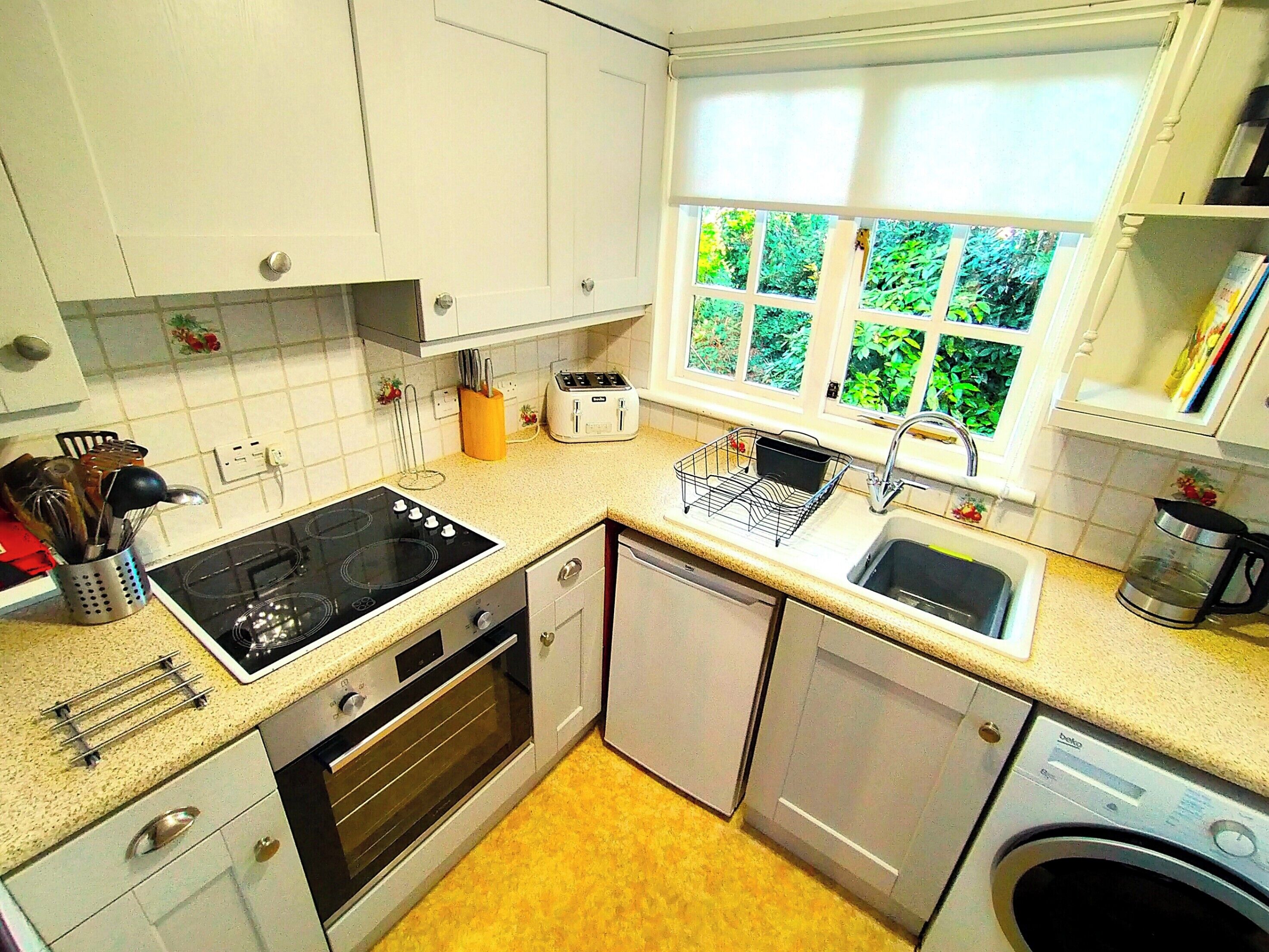 A compact kitchen with a stove, oven, dishwasher, and sink. There is a window with a view of greenery outside.