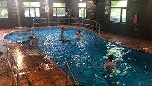 Sisätiloissa sijaitseva uima-allas, jossa useat ihmiset uivat ja leikkivät. Altaan ympärillä on ikkunoita ja turvallisuusmerkkejä.