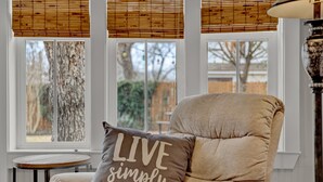 Ein gemütliches Wohnzimmer mit einem beigen Sessel, einem runden Holztisch und einem Fenster mit Blick auf Bäume und eine Hecke.