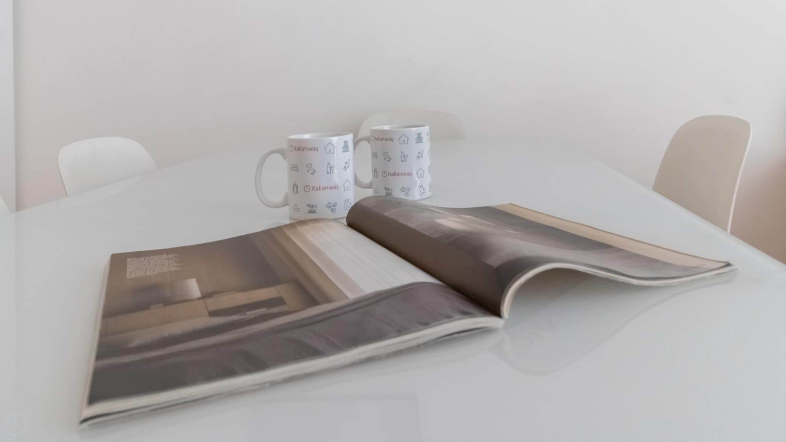 A table with a magazine, two mugs, and a chair.