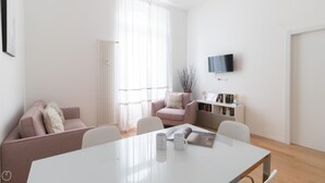 Ett modernt vardagsrum med ett vitt bord, en rosa soffa, en vit fåtölj och en bokhylla med böcker och dekorativa föremål.