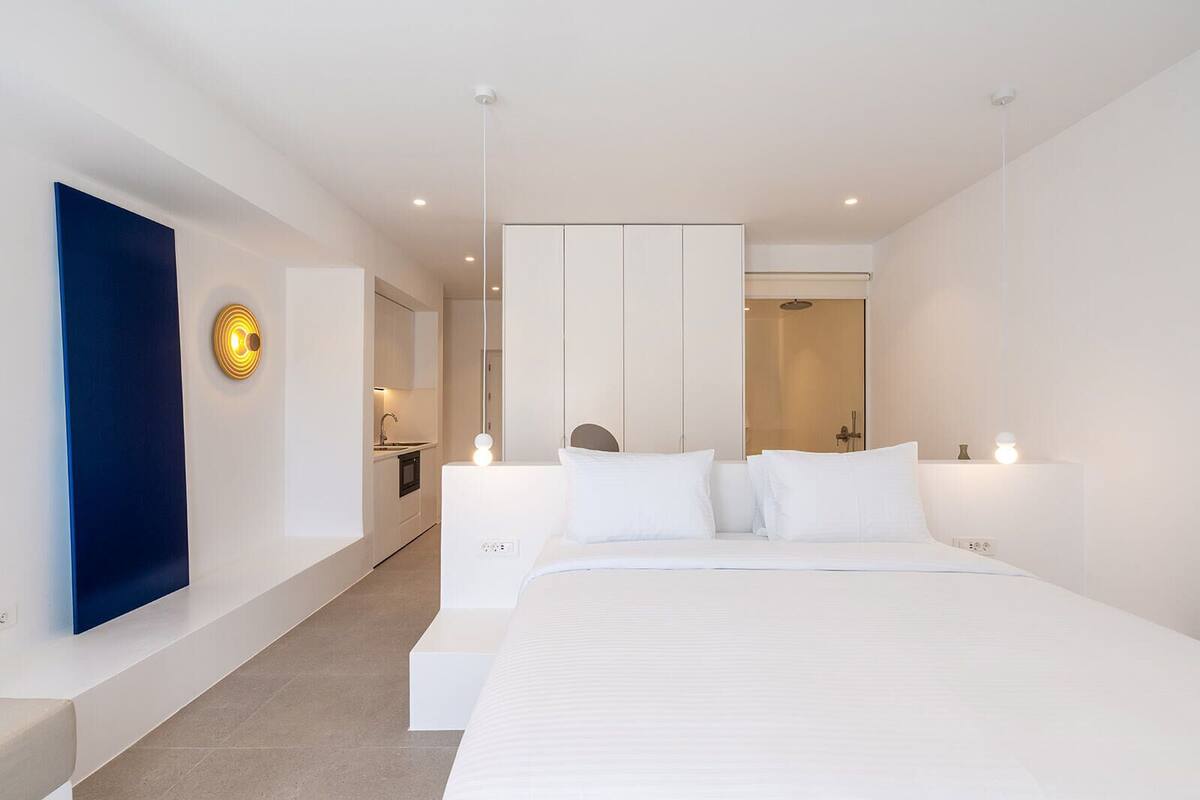 A modern hotel room with a large bed, a built-in wardrobe, a small kitchenette, and a bathroom with a glass shower.
