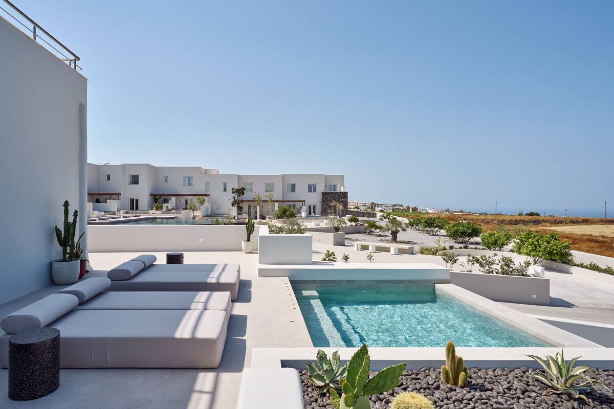 A modern outdoor pool area with a clear view of the sea, surrounded by white buildings and minimalist landscaping.