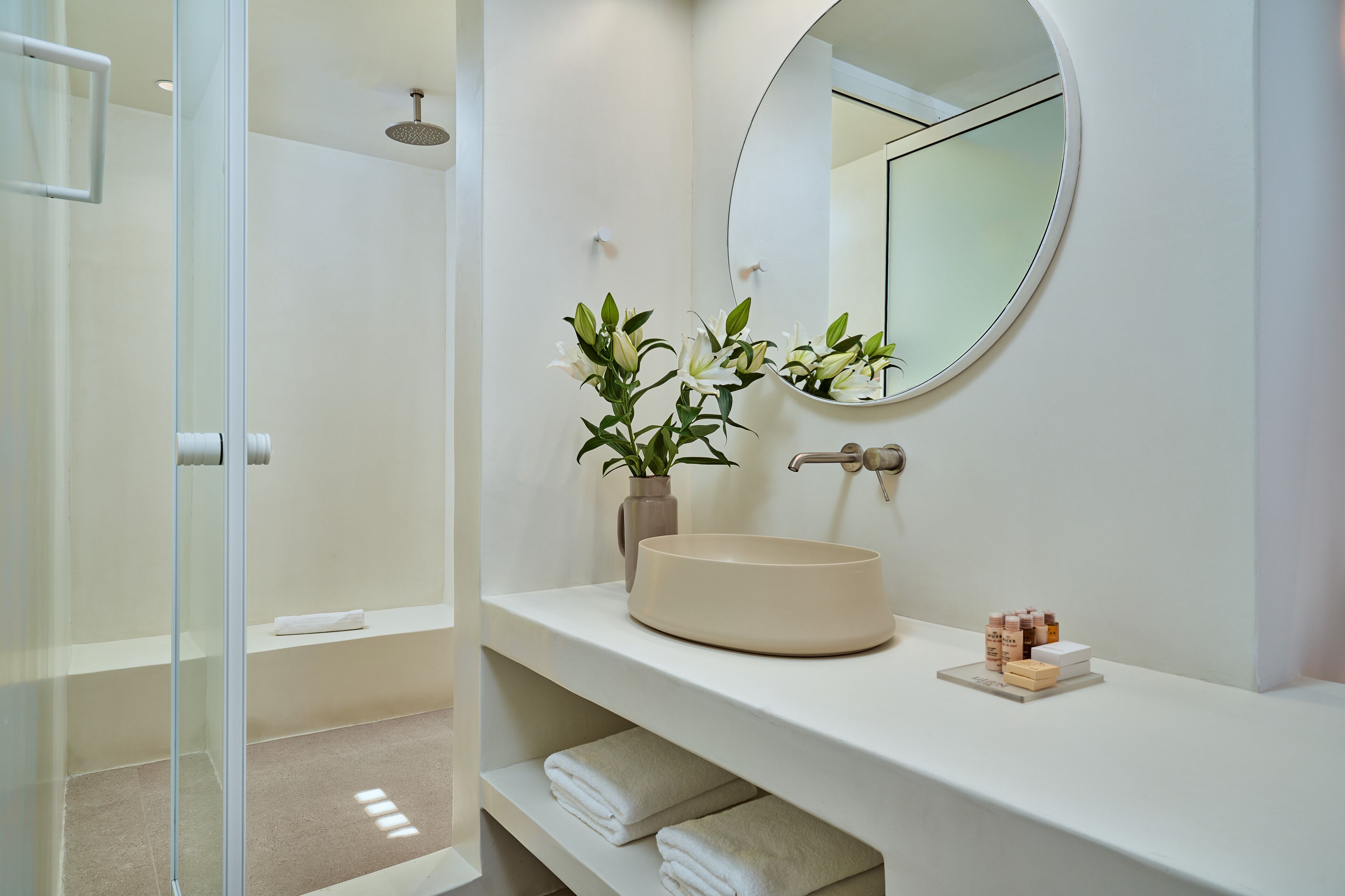 Un baño moderno con cabina de ducha de vidrio, un espejo redondo, un lavamanos color beige y un florero con flores blancas.
