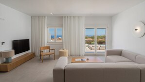 A modern living room with a large flat-screen TV, a beige sofa, a wooden coffee table, and a view of the outdoors through large windows. - Lilium Homes (Santorini)