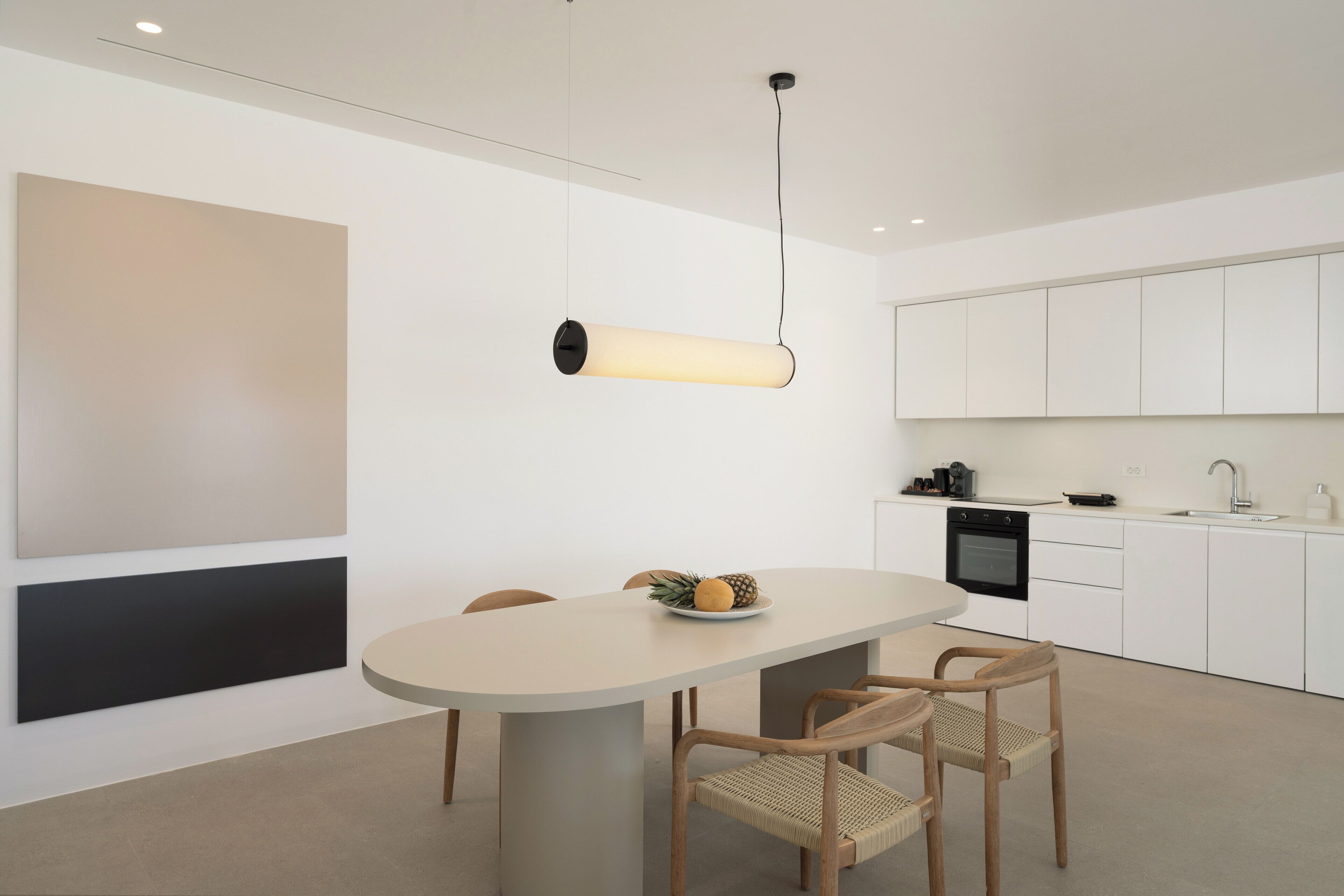 A modern kitchen with a round dining table, two wooden chairs, a hanging light fixture, and white cabinetry.