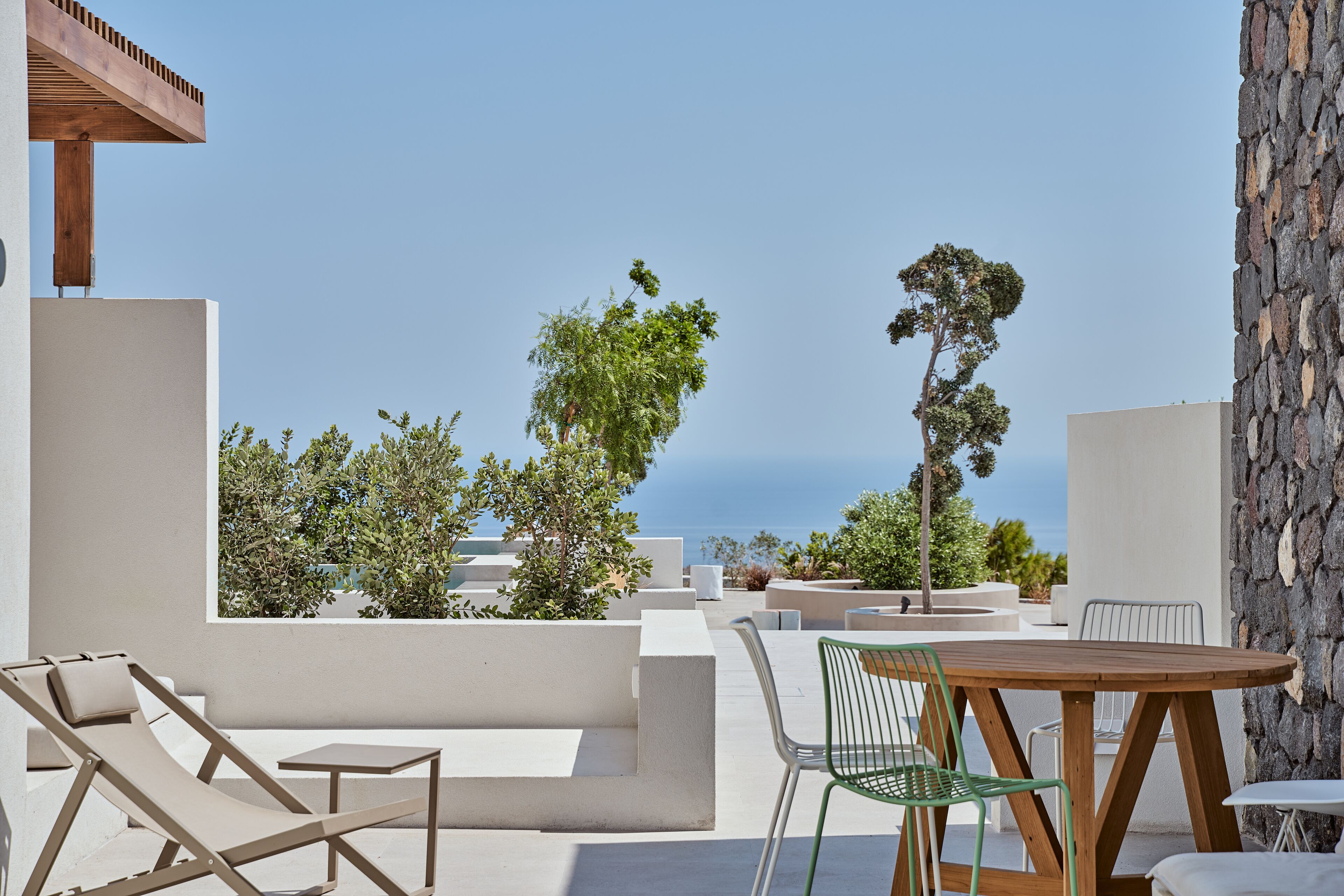 Un espacio moderno para sentarse al aire libre con una mesa y sillas de madera, rodeado de paredes blancas y follaje, con vistas a un cielo azul despejado y un paisaje lejano.