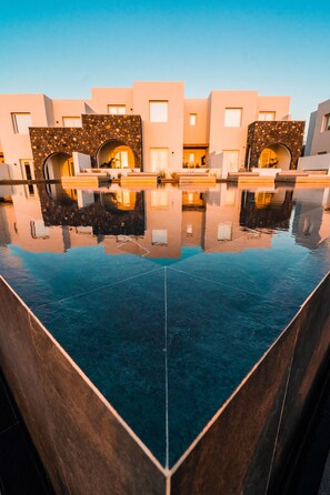 Un hotel moderno con una piscina reflectante, arcos de piedra y un cielo azul despejado.