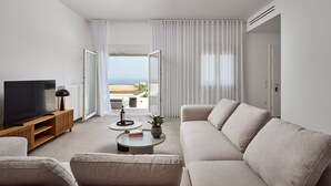 A modern living room with a large flat-screen TV, a sofa, a coffee table, and a view of the sea through large windows. - Lilium Homes (Santorini)