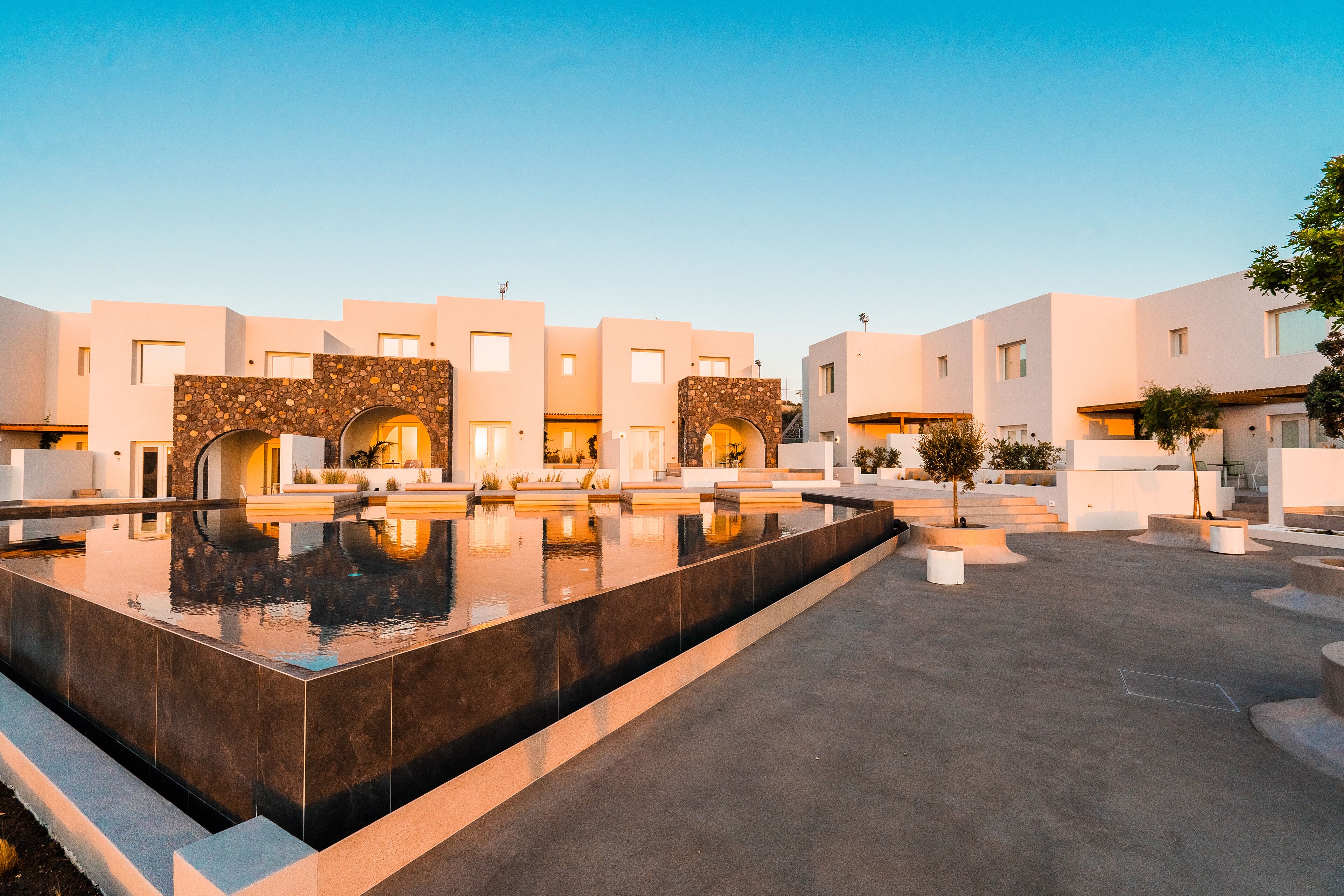 Un patio d’hôtel moderne avec une piscine centrale aux reflets changeants, des bâtiments blancs agrémentés d’éléments en pierre et des espaces de détente en plein air.