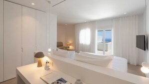 A modern hotel room with a large bed, a desk with a lamp, a bedside table with a phone, and a view of the sea through the window. - Lilium Homes (Santorini)