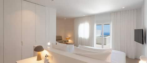 A modern hotel room with a large bed, a desk with a lamp, a bedside table with a phone, and a view of the sea through the window.