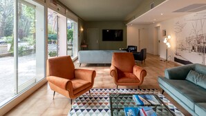 A spacious living room with large windows, a blue sofa, two orange armchairs, and a coffee table with magazines. - Hotel Millepini (Ravenna)