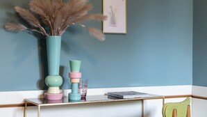 A corner room with a teal wall, a wooden chair with a green cushion, a white desk with a vase of pampas grass, and a framed picture. - Hotel Millepini (Ravenna)