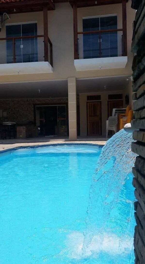 Una piscina con cascada, un edificio de dos pisos con balcones y un área de patio techada.