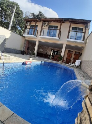Una piscina con cubierta de madera y sillas de descanso, un edificio de dos plantas con balcones y ventanas amplias, y una antena parabólica en el techo.