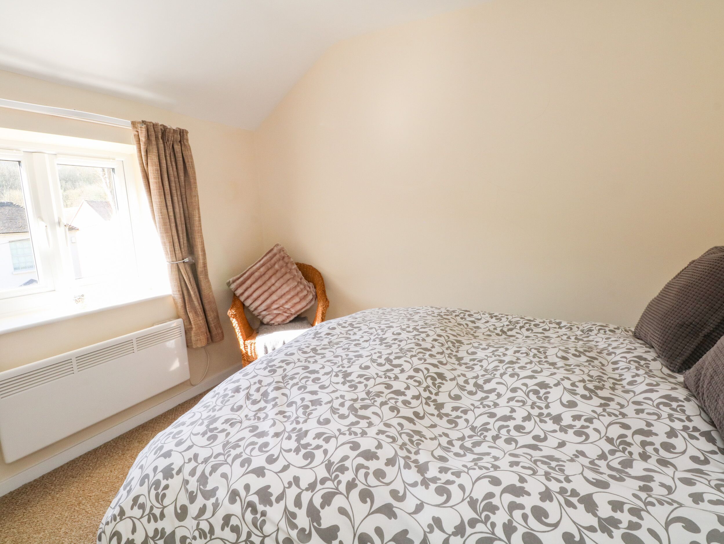 A bedroom with a large bed, a window with curtains, a radiator, and a chair with a cushion.