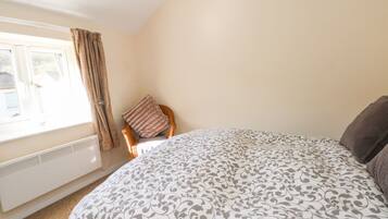 A bedroom with a large bed, a window with curtains, a radiator, and a chair with a cushion.