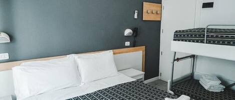 A hotel room with a single bed, a bunk bed, a bedside table, a lamp, and a wall-mounted phone.