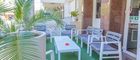 Un espacio al aire libre con sillas blancas y una mesa, rodeado de plantas en macetas y un suelo de césped artificial verde.