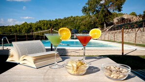 A poolside table with a book, two drinks with orange slices, and bowls of snacks. - Tenuta di Forte Sorgnano | Farmhouse with private pool (Gualdo Cattaneo (PG))