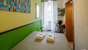 Una habitación con una cama, un ventilador de techo, un cuadro abstracto colorido, una ventana con cortinas y una pequeña mesa con una planta.
