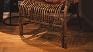 Una silla de mimbre con un plaid de piel y un cojín rojo, ubicada junto a una mesa con una lámpara.