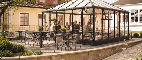A glass greenhouse structure with tables and chairs, surrounded by a stone wall and plants.